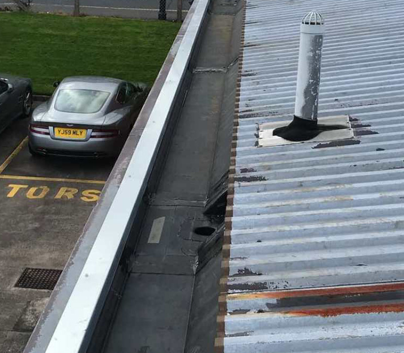 view of a rooftop with a white ventilation pipe and a gray sports car parked below on a lot marked for motors showcasing a gray building with weathered roofing suggesting the number seven through its lines and angles